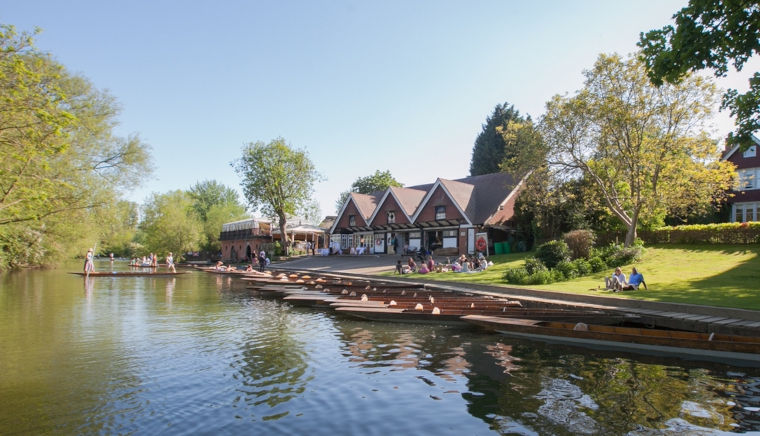The river Cherwell in spring sunlight with punts along the bank, students sitting on the lawn and the Cherwell Boathouse.