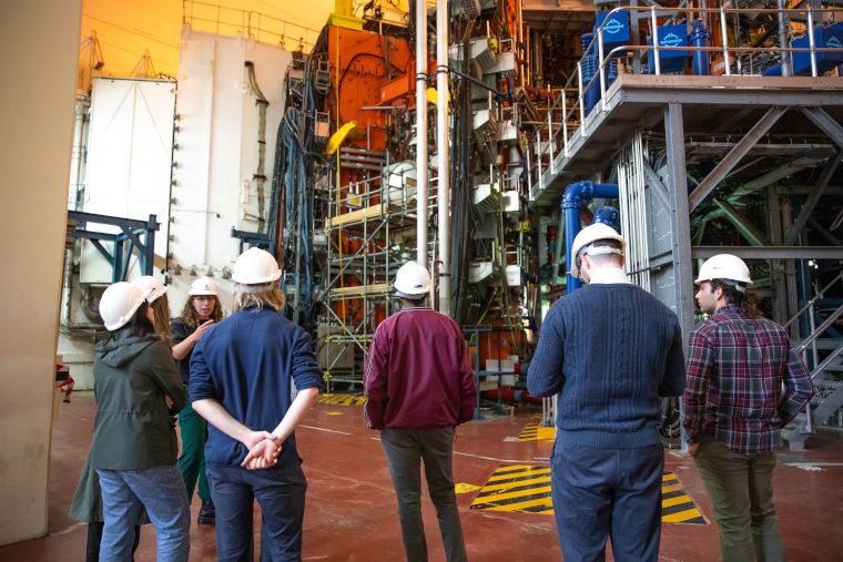 Members of the public wearing hard hats listen to an expert explaining a massive fusion energy machine at UKAEA's Culham Campus.