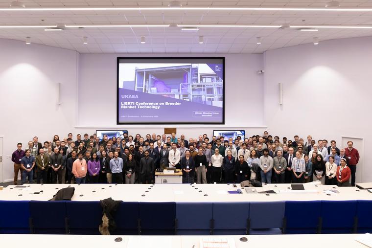 A large group of people attending UKAEA's LIBRTI Conference are facing the camera and stood in an auditorium Infront of a presentation.