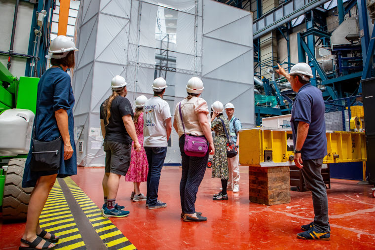 An expert from UKAEA shows visitors a fusion energy machine at Culham Campus. Everyone is wearing a white safety helmet.