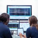 2 experts working on a problem in front of a screen with data on it.
