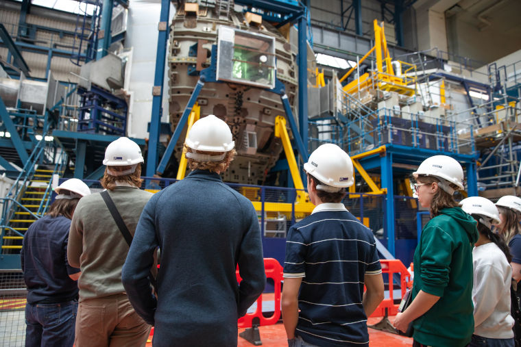 A group of visitors wearing white hard hats listen to an expert while looking at a fusion energy machine at the UK Atomic Energy Authority.