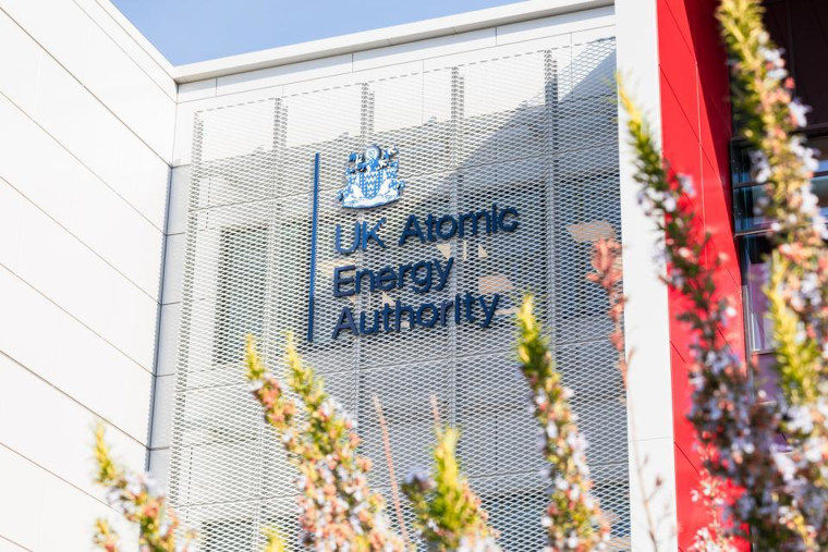 The UK Atomic Energy Authority crest and logo on the front of a large facility building through trees in blossom.