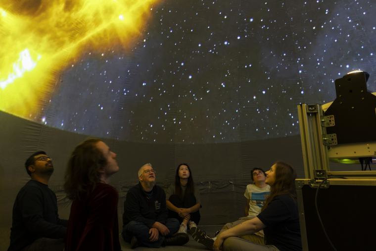 Five people site in the UKAEA Sun Dome. Their faces are highlighted by the large projected image of the Sun and stars above them.